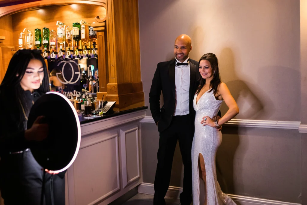 Roaming photo booth operator uses a ring light to photograph a well-dressed couple posing beside a bar at an indoor event, with liquor bottles and warm lighting in the background.