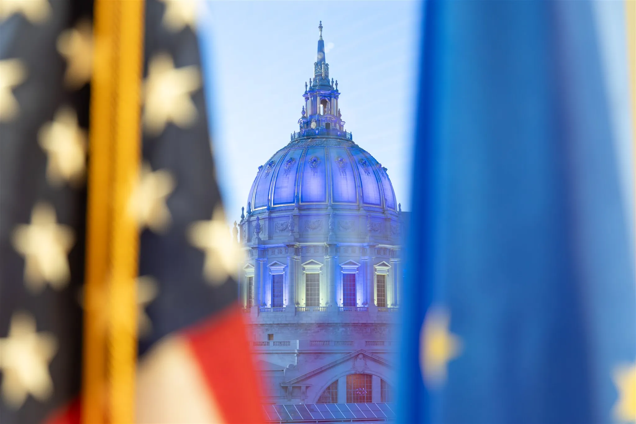 San Francisco City Hall illuminated in blue, framed by out-of-focus American and European Union flags in the foreground.