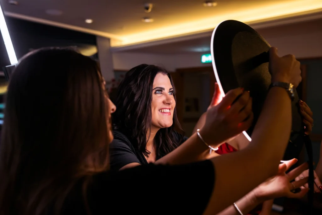 Two women at an indoor event smile as one adjusts a hat on another person, illuminated by a glowing ring light, creating a warm, candid moment.